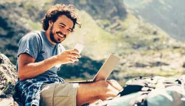 man sitting on rocks with phone and laptop