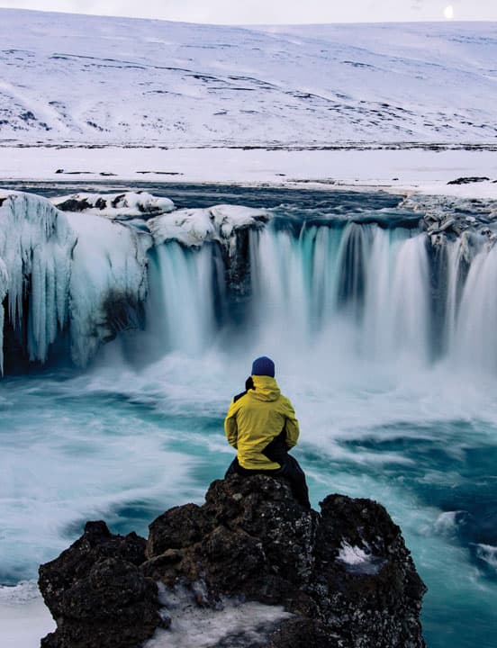 person on rock in front of waterfall in winter